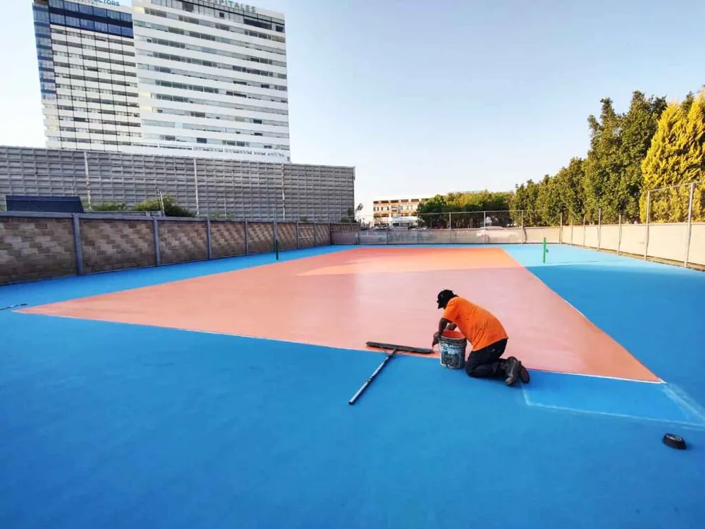 Aplicación de pintura acrílica con jalador sobre una cancha deportiva en azul y naranja durante la restauración de canchas de basquetbol y tenis